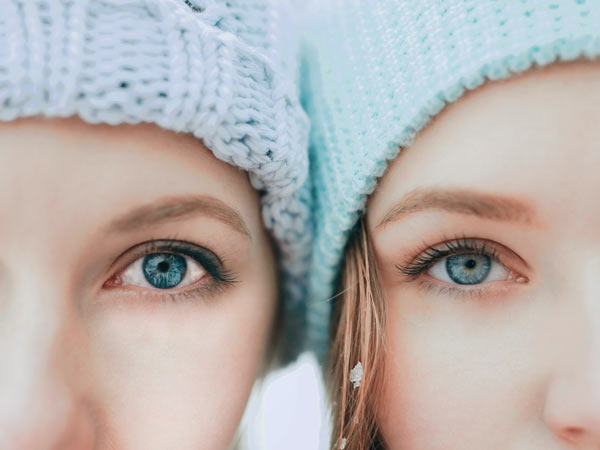 دو خانم کنار یکدیگر ایستاده اند. هر دو چشما هایی رنگی دارند و کلاه بافتنی به سر کرده اند.