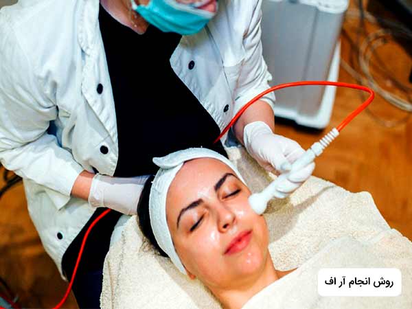يک خانم با چشمان بسته روي تخت دراز کشيده است. پزشک در حال زيباسازي پوست بدن او با استفاده از روش راديوفرکانس درماني مي باشد.خانم لباس سفيد به تن دارد